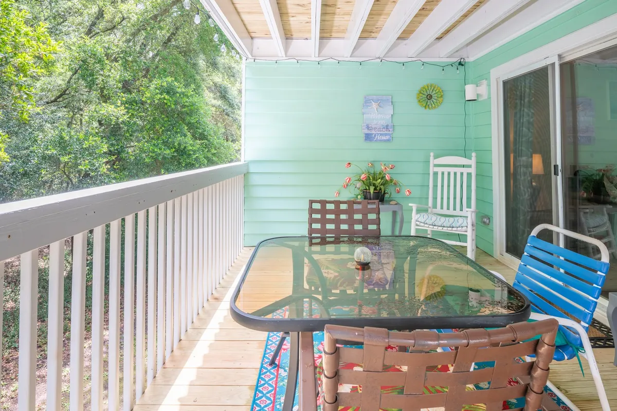 A cozy outdoor dining area on a porch or balcony, featuring a glass dining table surrounded by colorful chairs, lush potted plants, and string lights hanging overhead. The setting is perfect for sunset drinks or a quiet meal while enjoying the outdoors.
