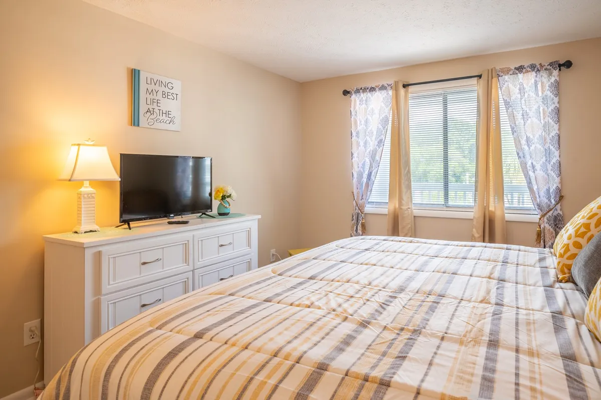 A tranquil bedroom featuring a bed with striped bedding, a flat screen TV on a dresser, a window with curtains letting in soft light, and tasteful decorative wall art. The room feels cozy, inviting, and ideal for relaxation or enjoying a quiet evening in.