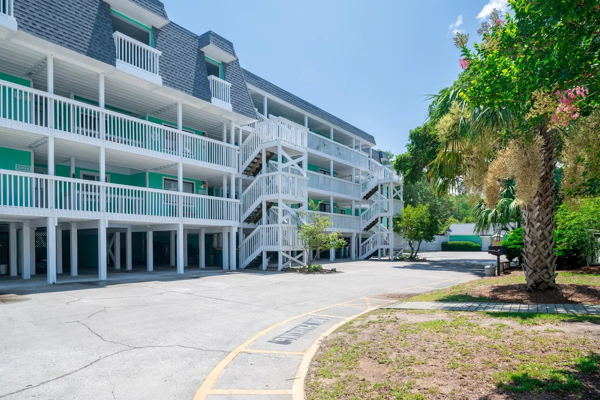 This image features the exterior of a three-story building with balconies, surrounded by lush greenery and a paved parking area in the foreground. Guests can be seen walking around the property, enjoying the garden, and socializing on the balconies.