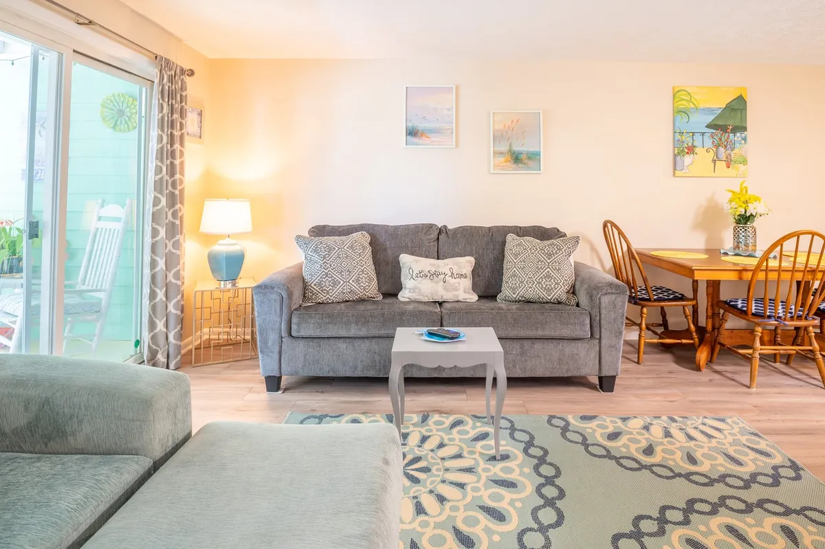 An inviting open-plan area showcasing a plush sofa, a dining table set for six, a soft rug, lamp, and coastal-inspired artwork. The space is designed for both intimate dinners and lively game nights, with a cohesive modern-coastal style.