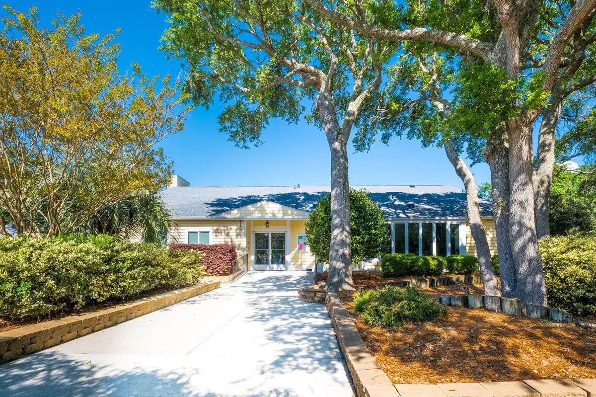 The image features the building's front entrance framed by large trees and manicured landscaping. The entry pathway invites guests toward the door, with soft natural light and greenery creating an immediate sense of arrival and tranquility.