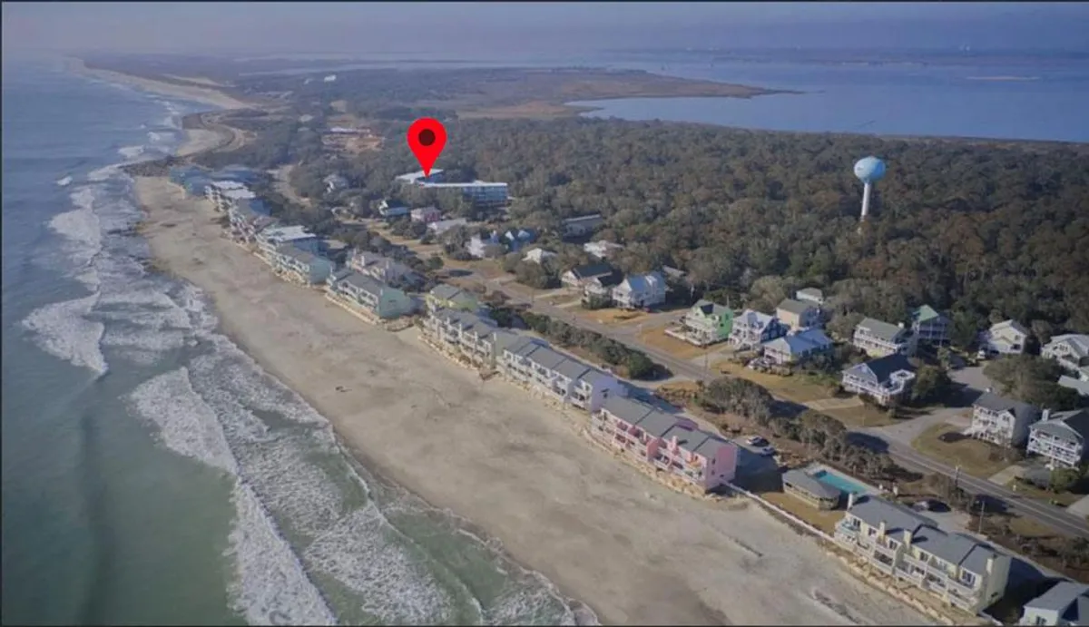 A stunning exterior shot of an uncrowded sandy beach, with gentle ocean waves lapping the shore and colorful beach houses nestled among lush greenery. The water tower in the background adds a local touch, and the overall mood is serene and romantic.