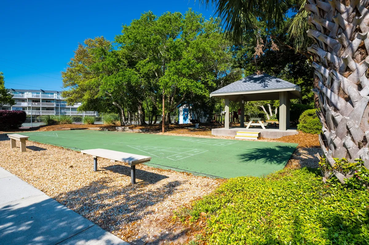 A cheerful outdoor area featuring a green playing surface, a white gazebo, benches, and surrounding trees. The space is set up for games, family gatherings, or relaxed picnics, inviting children and adults to enjoy the outdoors together.