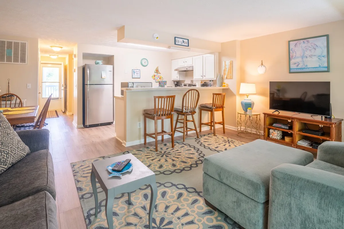 The image showcases a bright, open living space with a comfortable sofa facing a large TV, a sleek kitchen counter with bar seating, and a cozy dining area. The layout is thoughtfully designed for both relaxation and togetherness, with tasteful beach-inspired decor enhancing the welcoming mood.