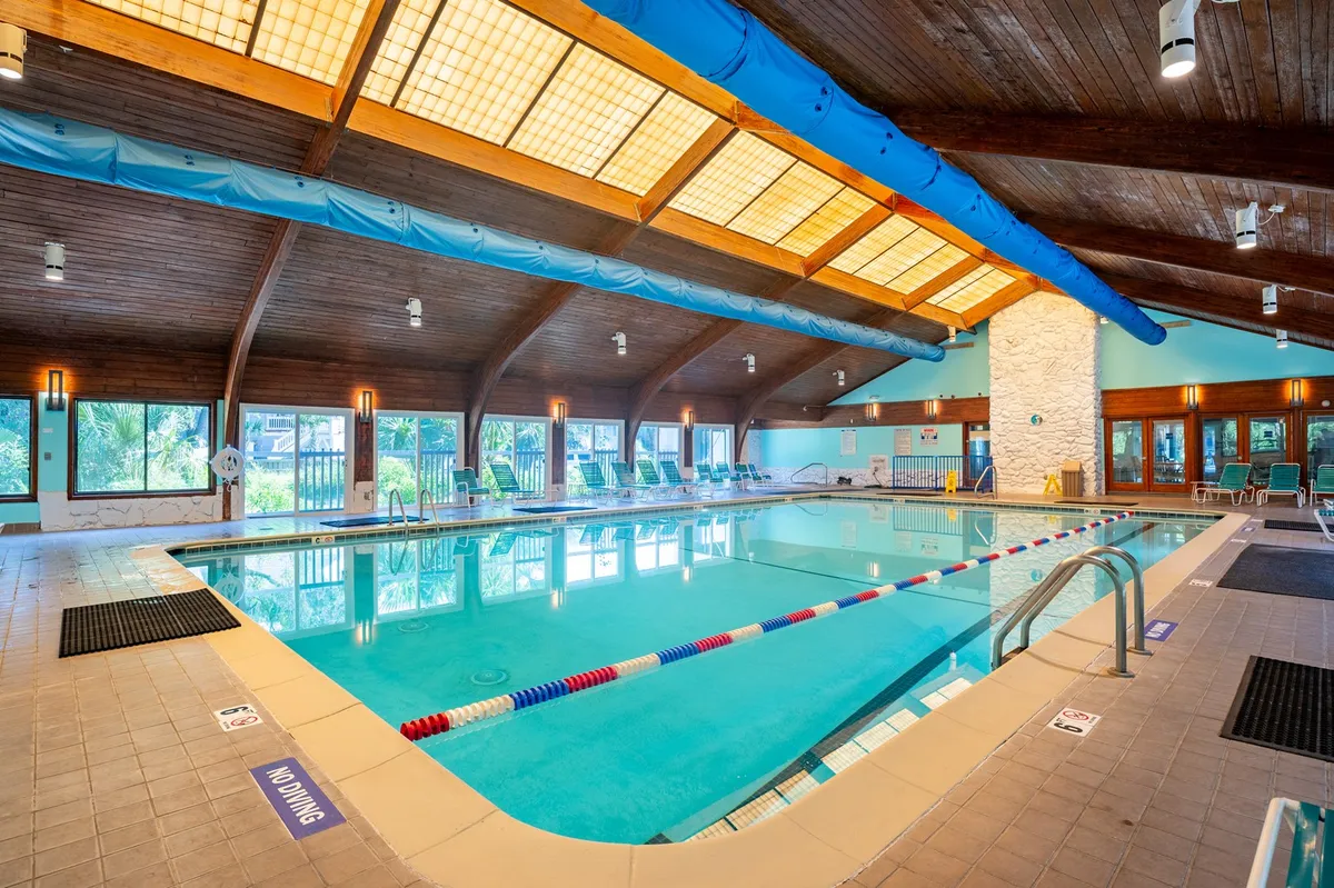 This image displays an indoor swimming pool with a wooden ceiling, large windows letting in natural light, and poolside loungers along the deck. The atmosphere is refreshing and relaxing, ideal for year-round recreation regardless of weather.