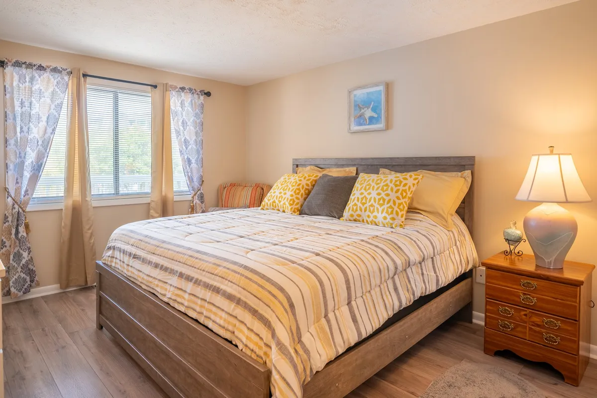 A comfortable and serene bedroom space, showcasing a plush bed with decorative pillows, a bedside lamp, and a window allowing natural light to gently illuminate the room. The space is styled for coziness and tranquility, perfect for unwinding or reading.
