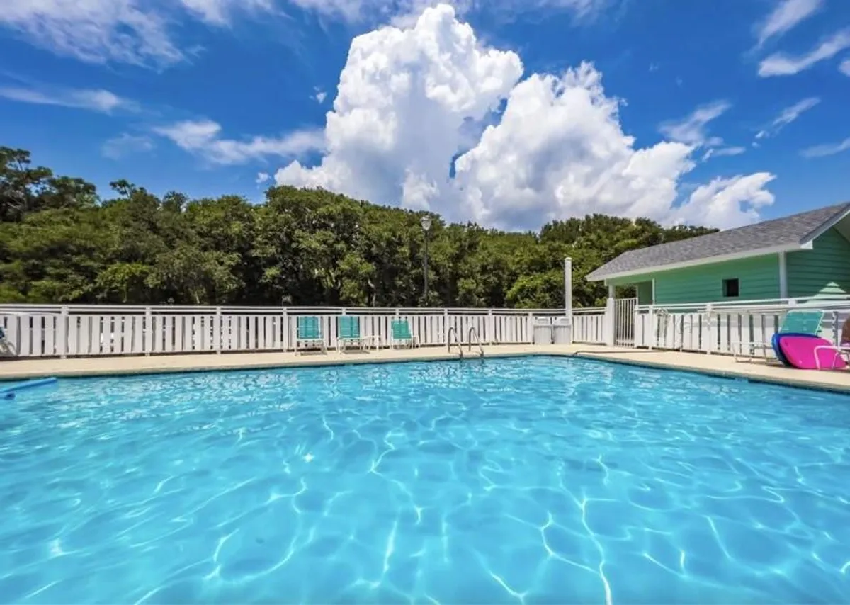 The scene centers on a sparkling outdoor swimming pool surrounded by lush greenery, with colorful lounge chairs inviting guests to swim, sunbathe, or simply unwind. The mood is leisurely and captures the essence of vacation relaxation.